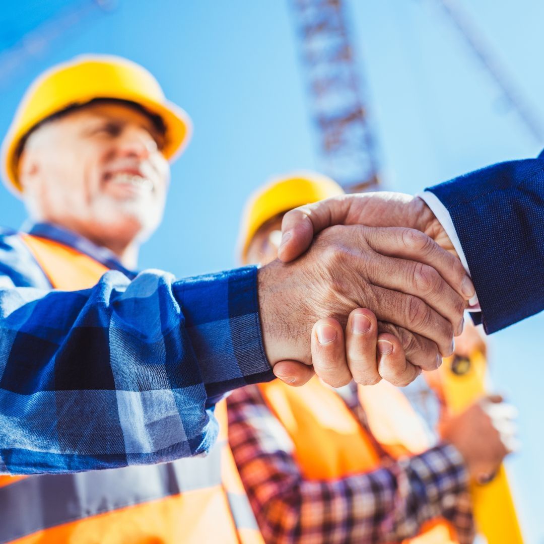 Zwei Personen mit Schutzhelmen reichen sich auf einer Baustelle die Hand